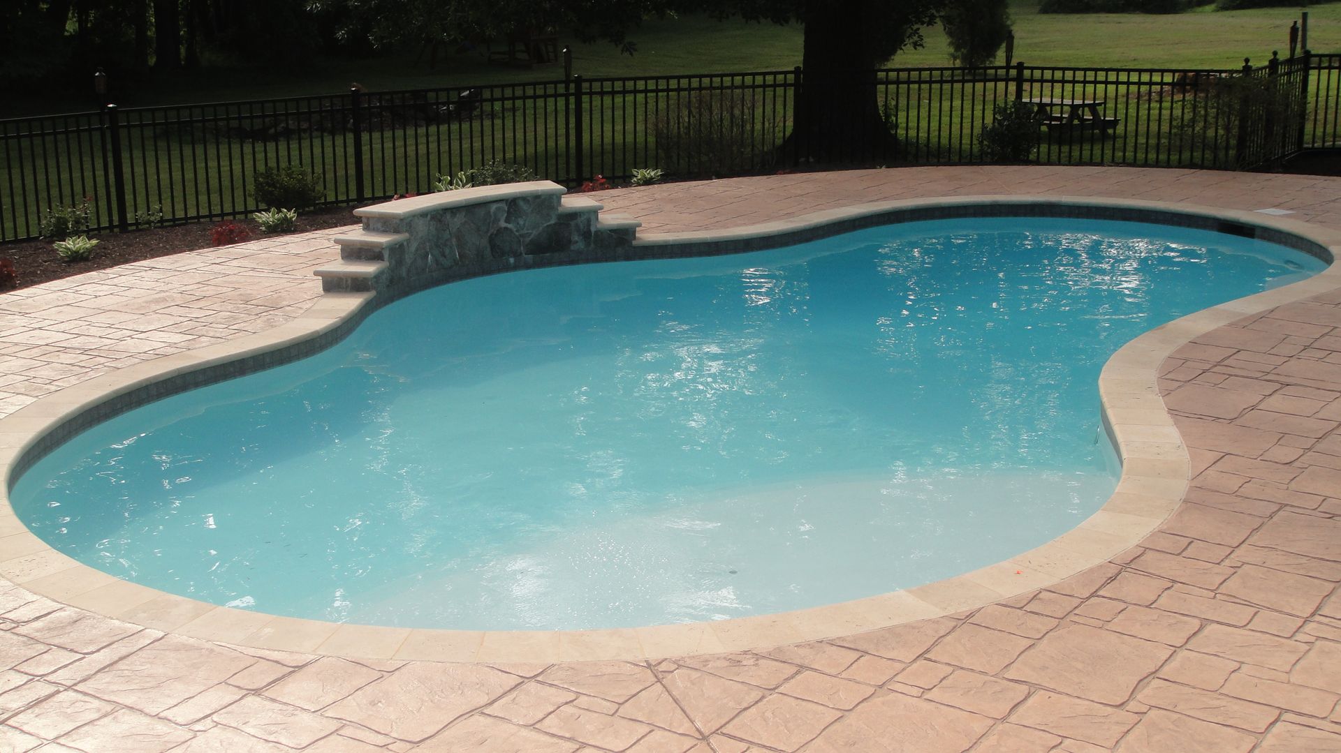Swimming pool with curved edges, surrounded by brick patio and black fence.