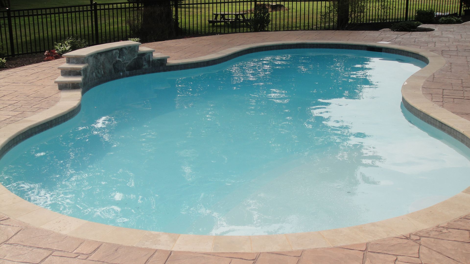A swimming pool with clear blue water and stone steps and border. Surrounded by patio and fence.