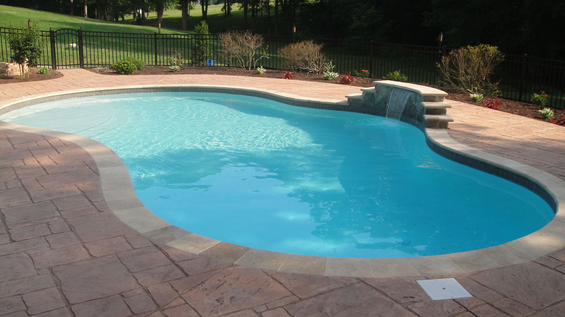 Swimming pool with light blue water and stone patio. Waterfall feature and black fence in the background.