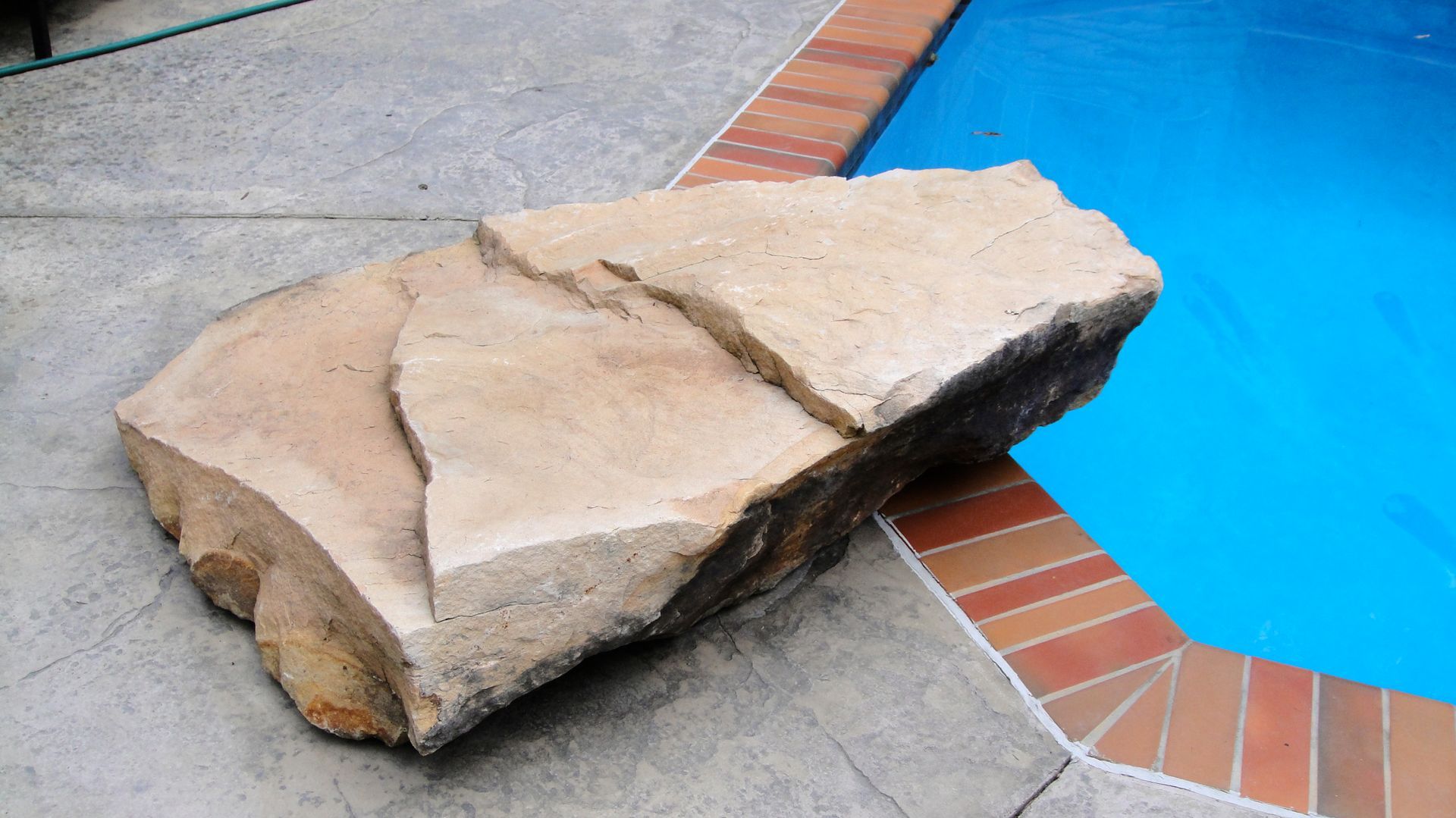 Large rock next to a pool. Brown rock on gray concrete, with blue water and brick edging.