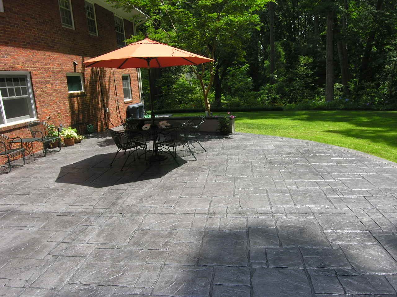 Brick house with gray concrete patio, orange umbrella, and wrought iron furniture on a sunny day.