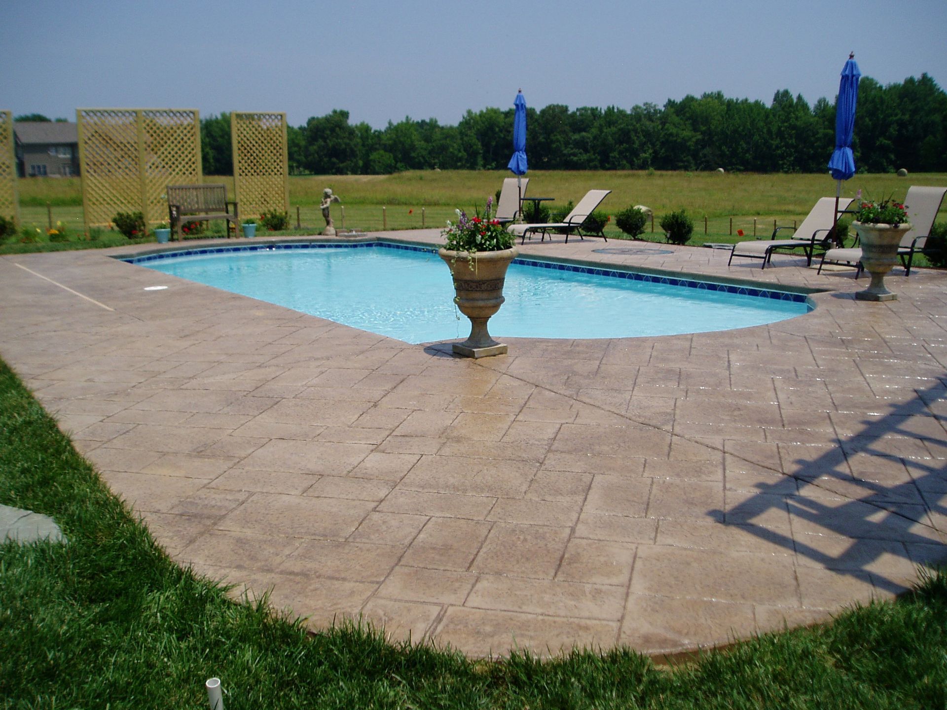 Pool with stamped concrete deck, lounge chairs, blue umbrellas, and a grassy lawn.