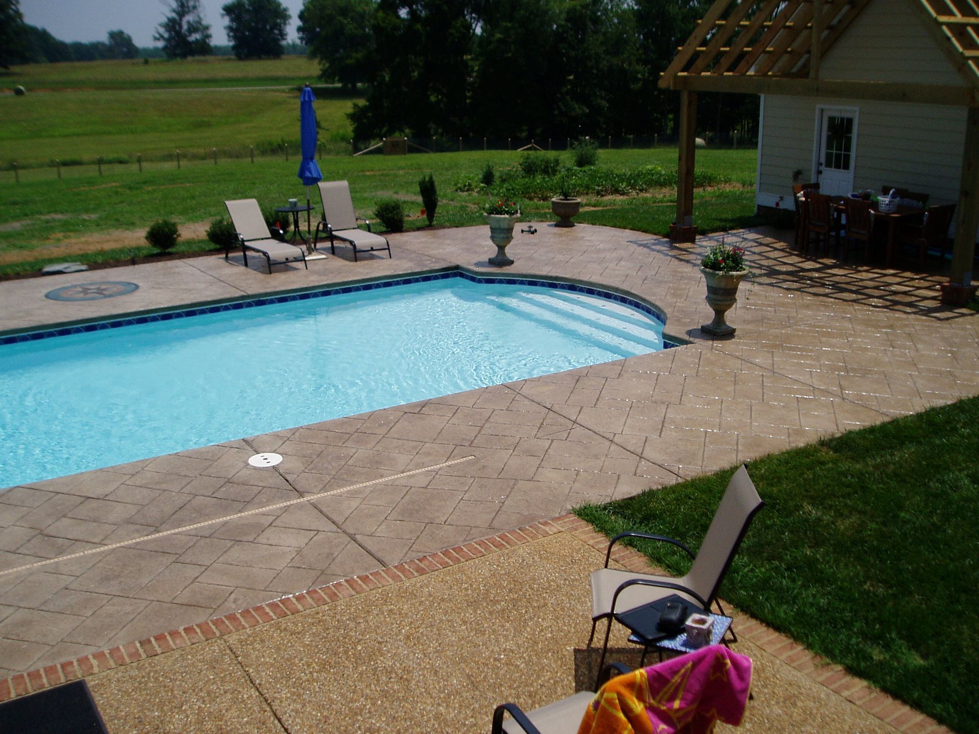 Poolside patio with pool, lounge chairs, small building, and grassy field.