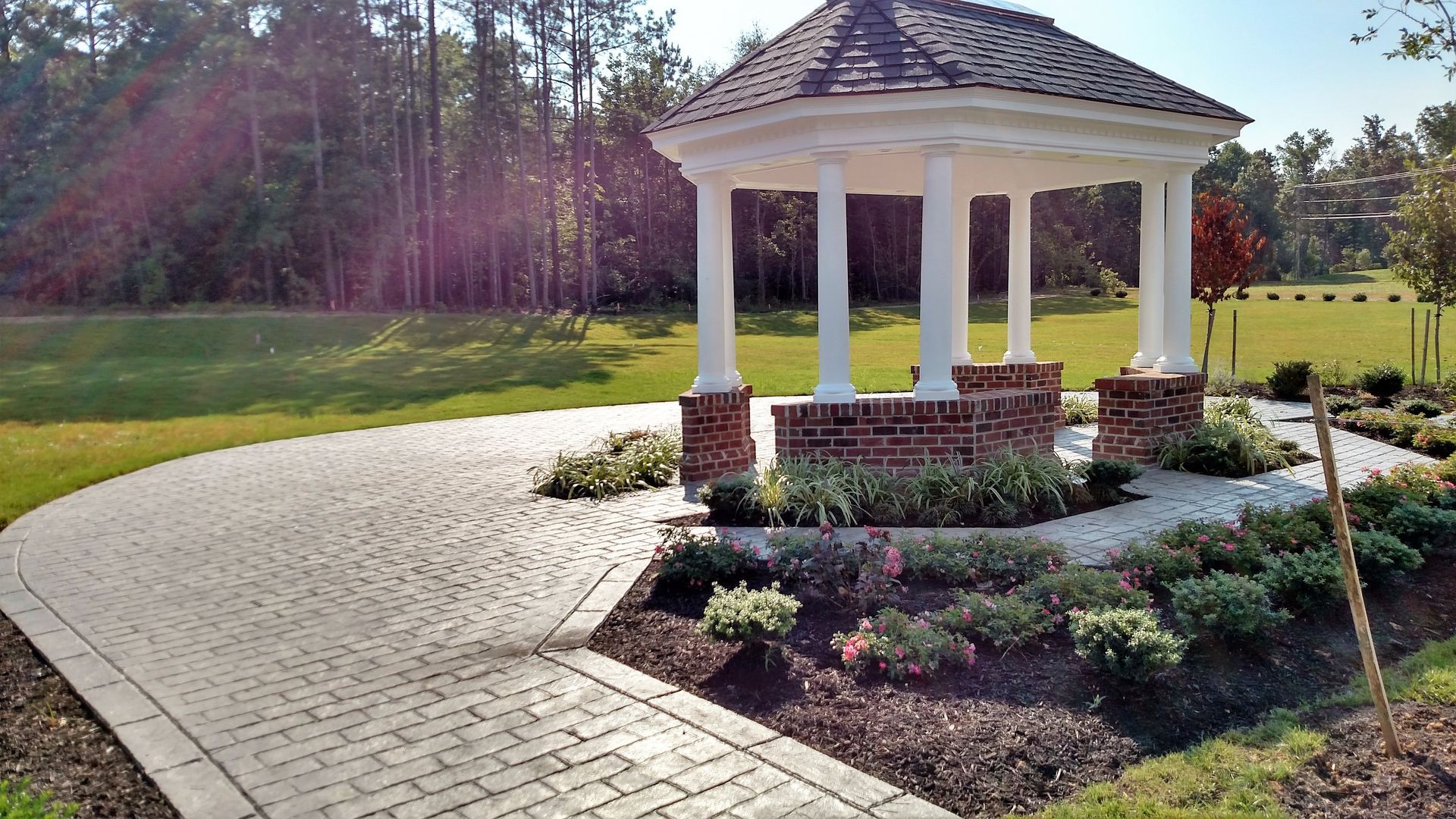 Gazebo with brick base, white columns, and brown roof surrounded by a garden and paved path.