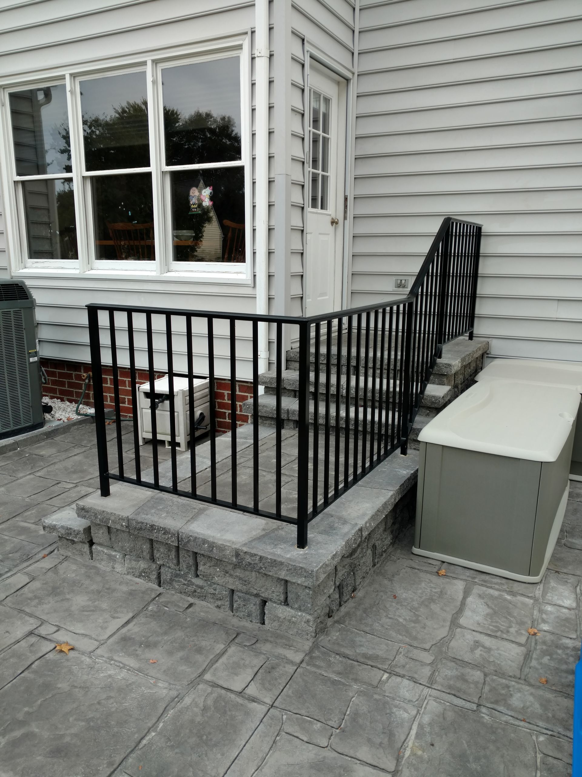 Black metal railing on a brick step leads to a door on a light gray house.