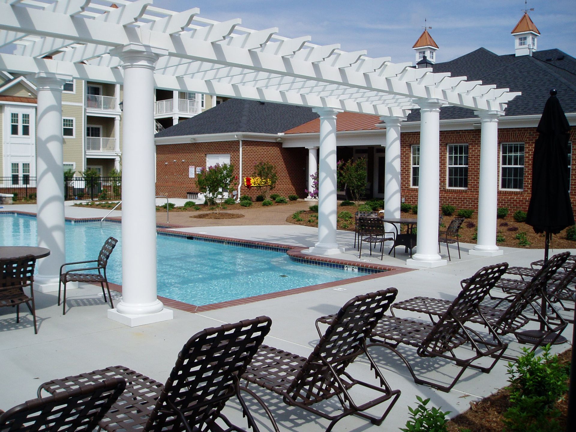 Pool area with white pergola, pool, lounge chairs, and building with red brick.