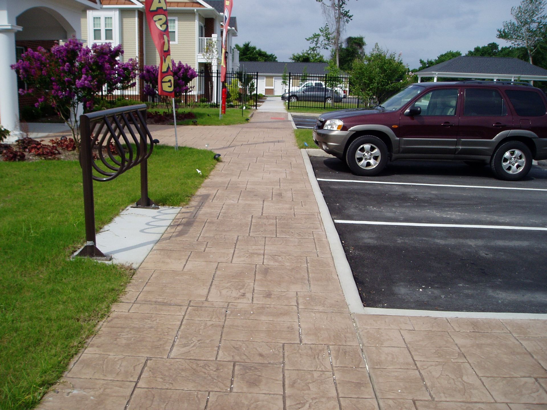 Sidewalk, grassy area, and parked SUV. Brown-stamped concrete sidewalk leads past a bike rack and a maroon SUV.