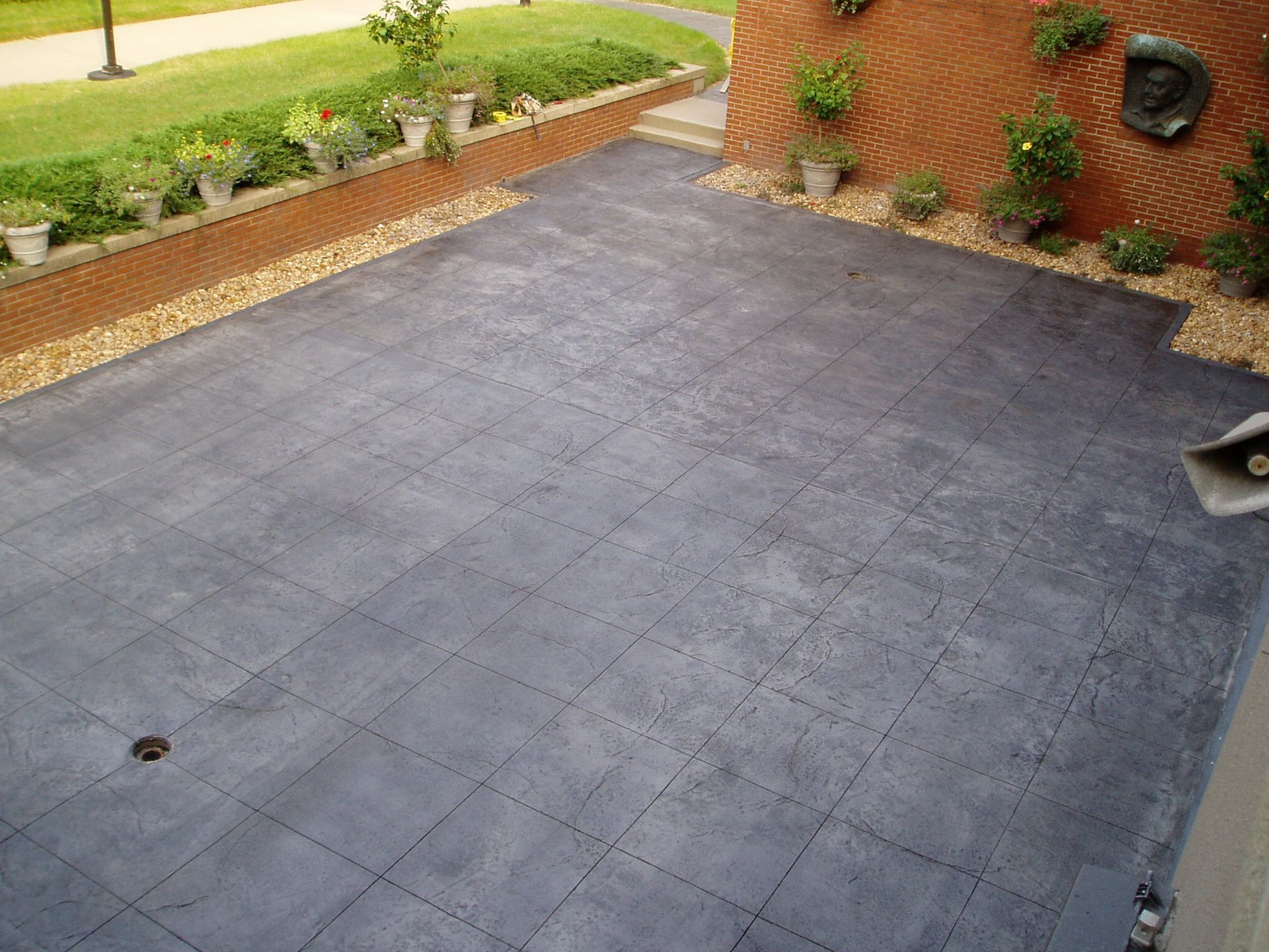 Brick patio with dark gray tiles, plants in pots along brick walls, grass and small rocks.