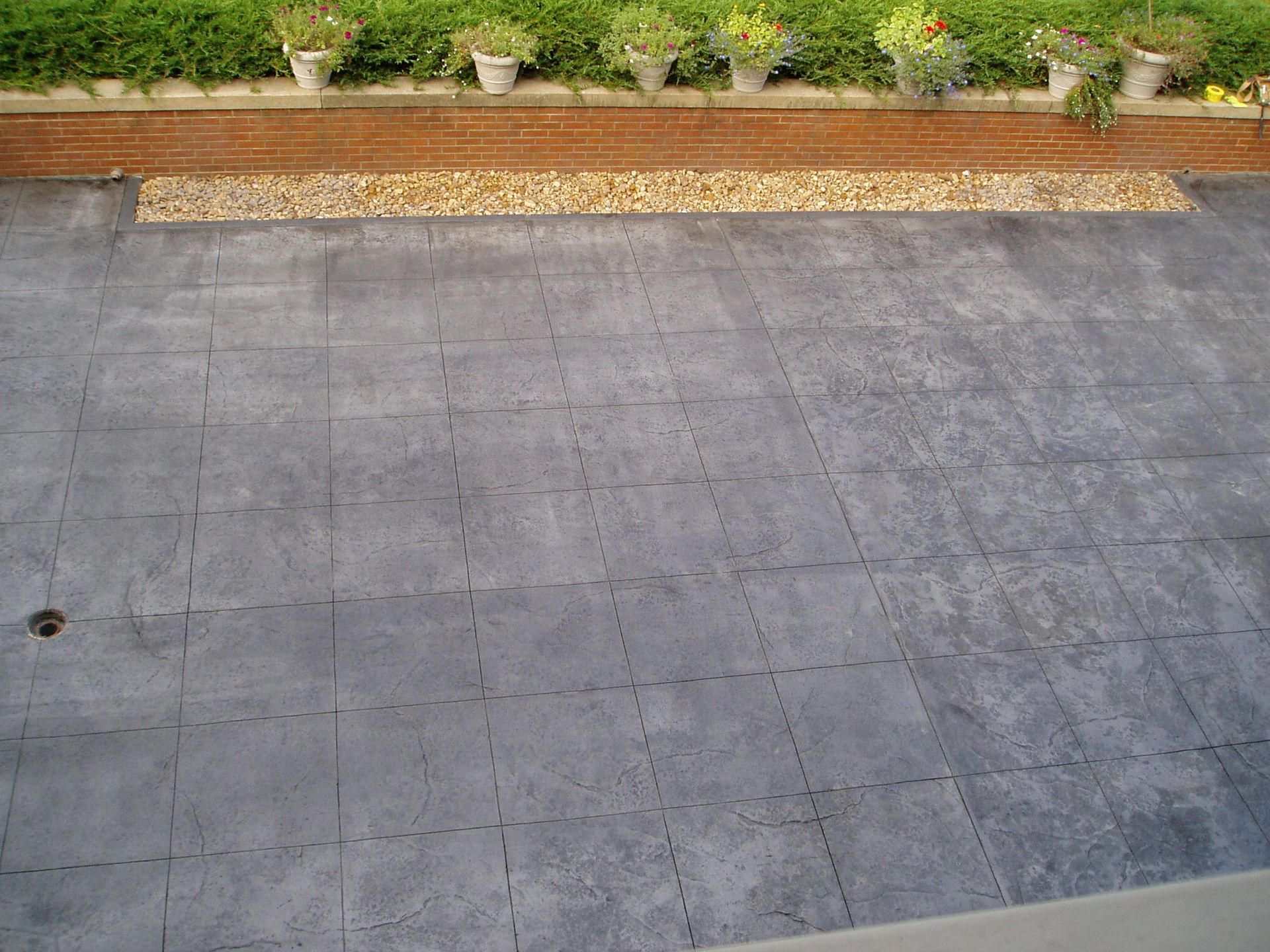 Gray tiled patio with a border of small rocks and a brick wall, green plants and pots in the background.