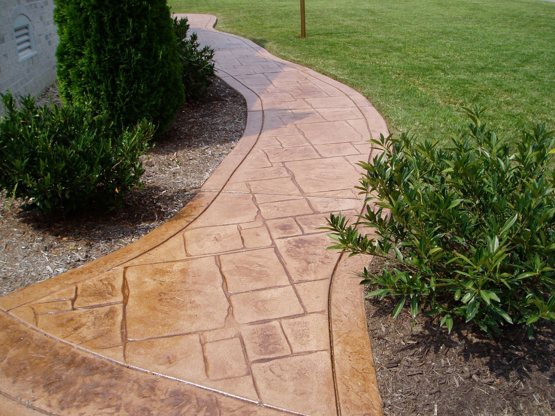 Stone-textured, curved walkway with brown edges, bordered by green grass, bushes, and trees.