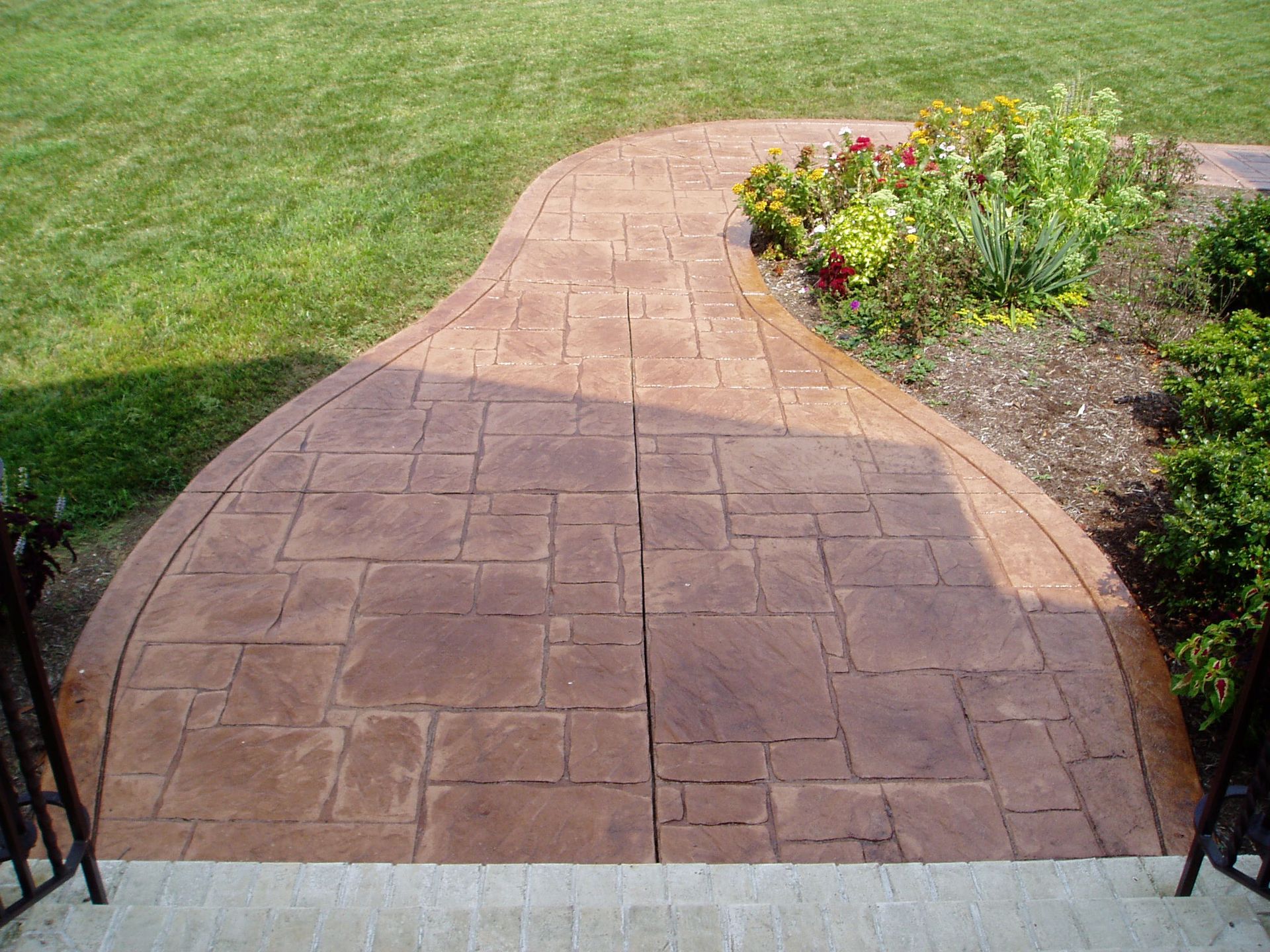 Brown stamped concrete pathway leading to a flower bed, bordered by green grass.