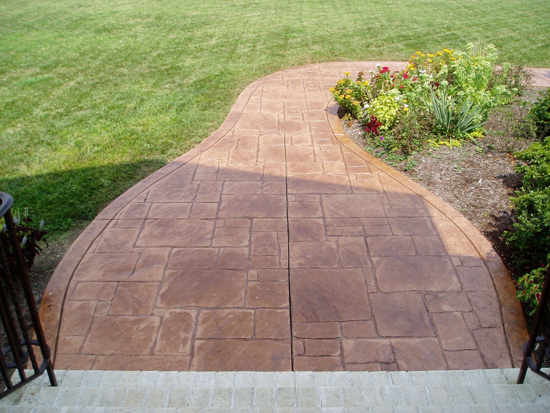 Brown stamped concrete pathway curves through a green lawn, leading to a garden.