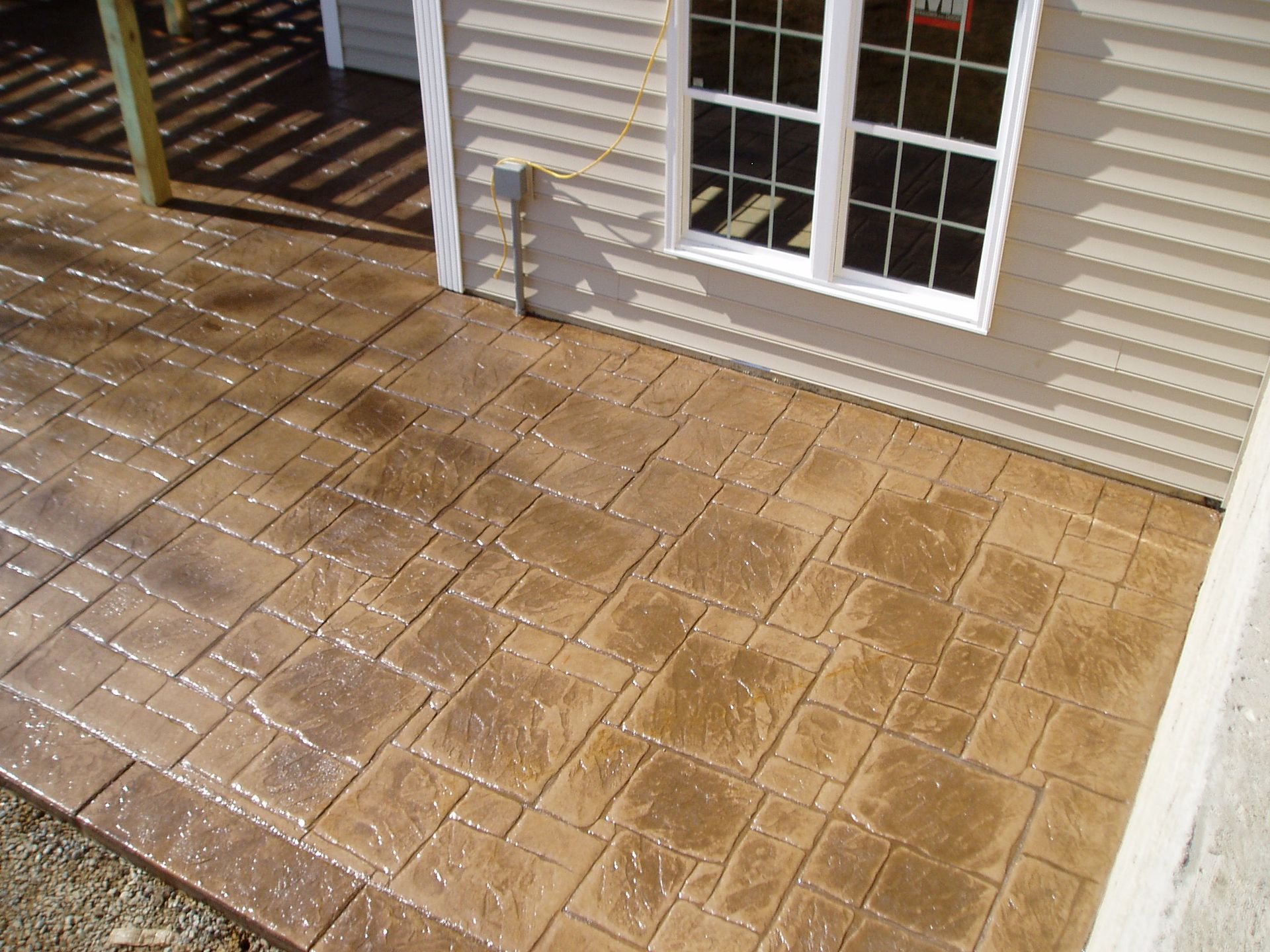 Stamped concrete patio with a light brown, rectangular pattern next to a house with a white window.