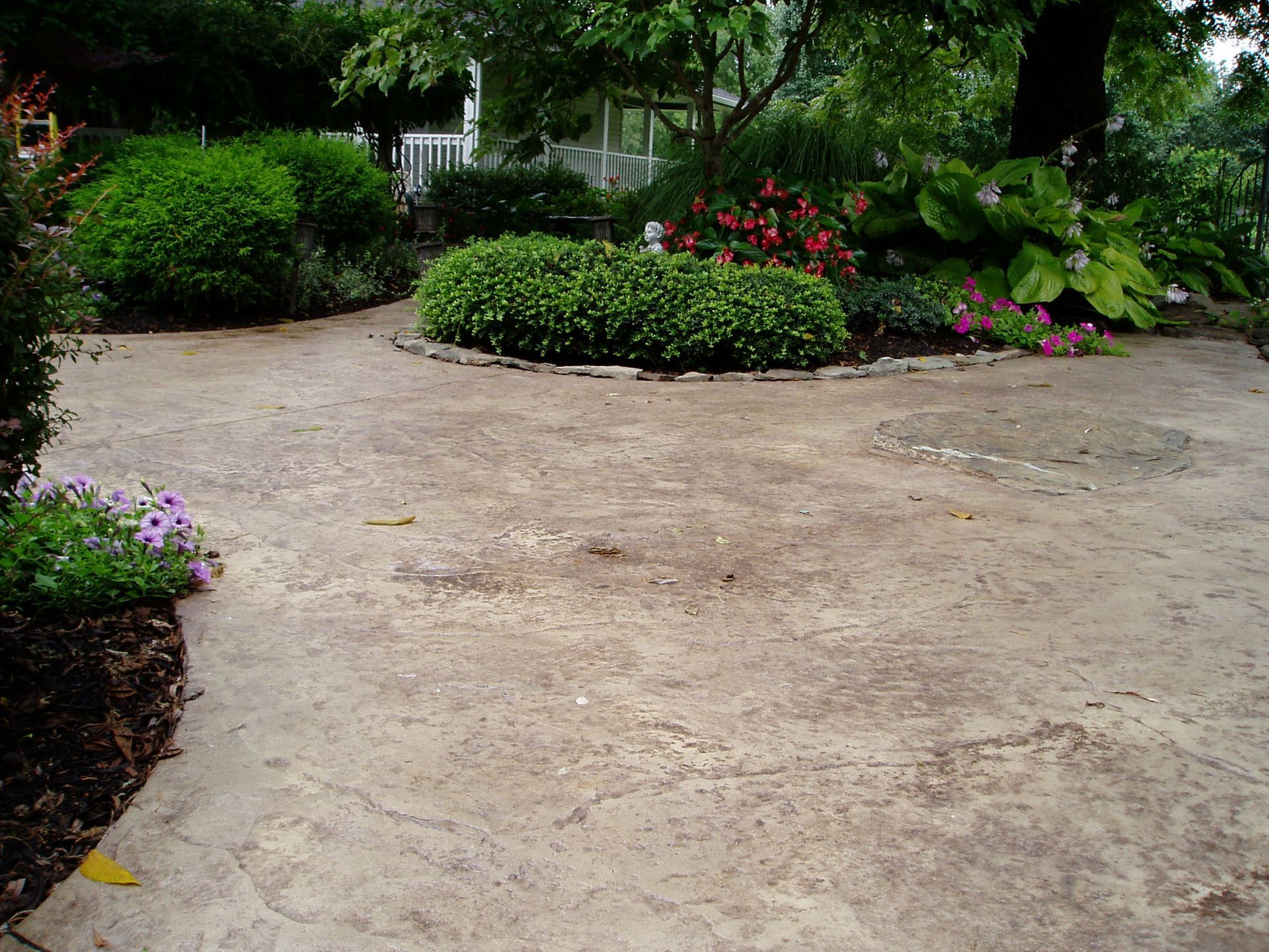 Stone patio with garden beds, featuring green shrubs and colorful flowers.