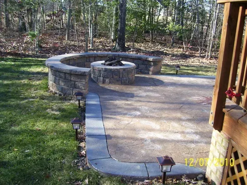 Stone fire pit and seating area on a concrete patio, surrounded by grass and trees.