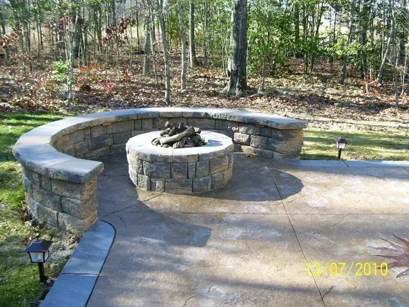 Stone fire pit with curved stone seating area on a concrete patio, surrounded by trees.