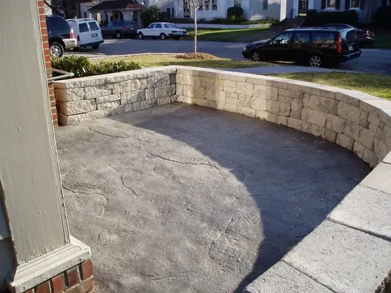 Curved, textured concrete patio enclosed by a stone retaining wall; cars and houses in background.