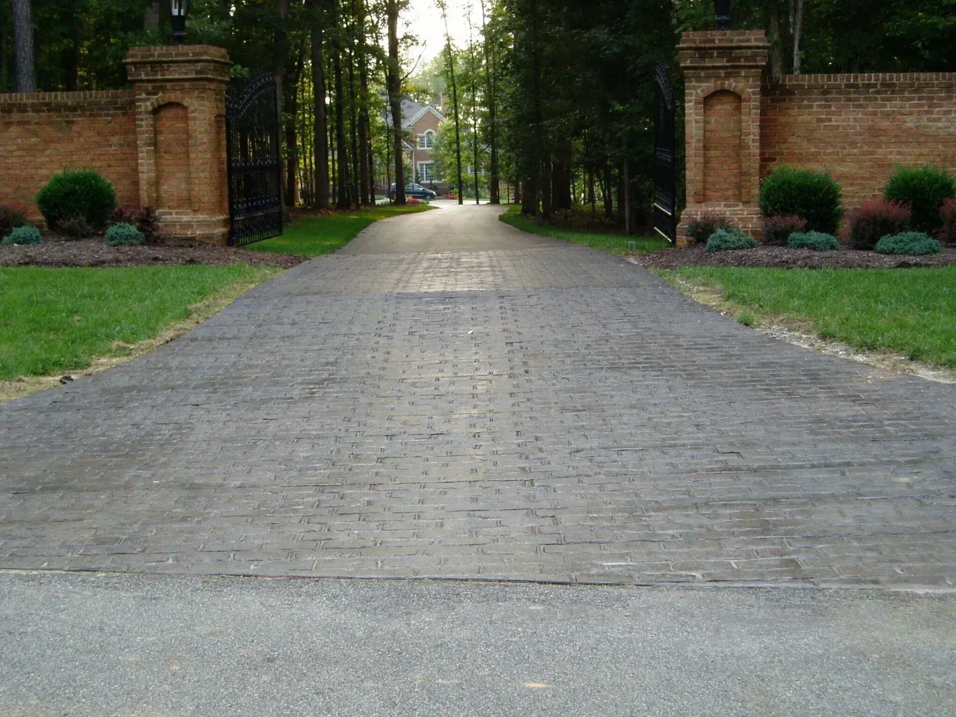 Driveway leading through brick gate posts towards a house, with trees on both sides and green lawn.