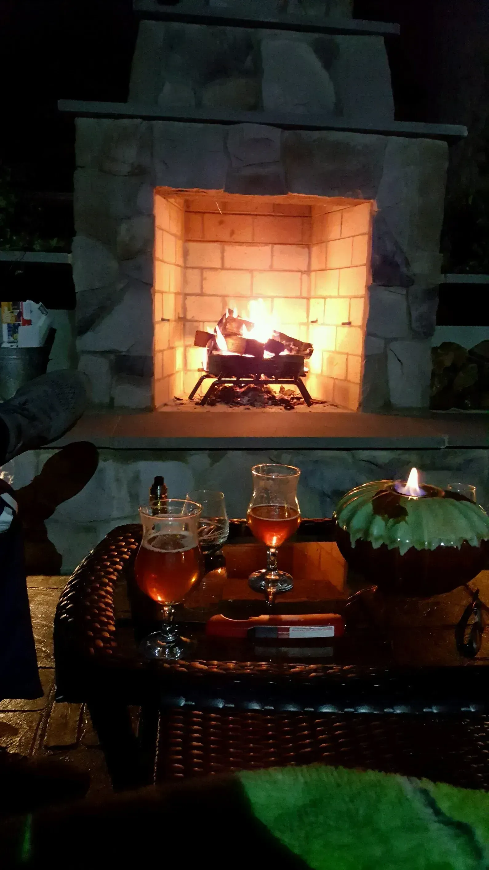 Lit outdoor fireplace with drinks and candle on a table at night.