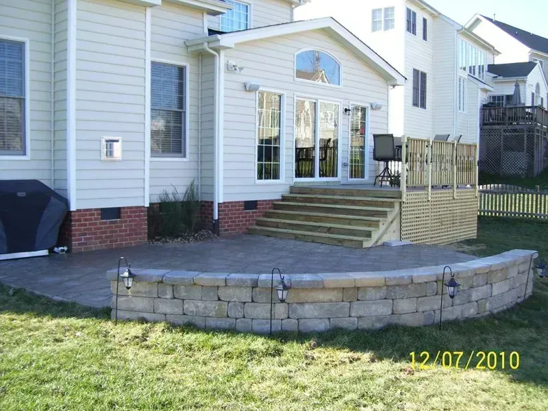 Backyard patio with stone retaining wall, steps to deck, and house in background.