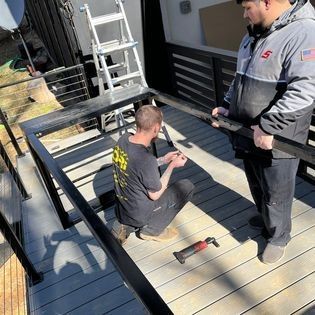 Two men are working on a deck with a ladder in the background.