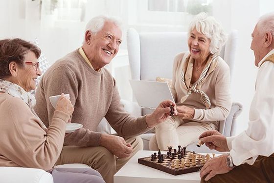 Four seniors are smiling, playing chess indoors, and a woman holds a teacup.