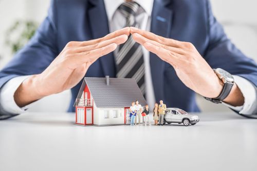Businessman shielding a miniature house, family, and car with hands over them