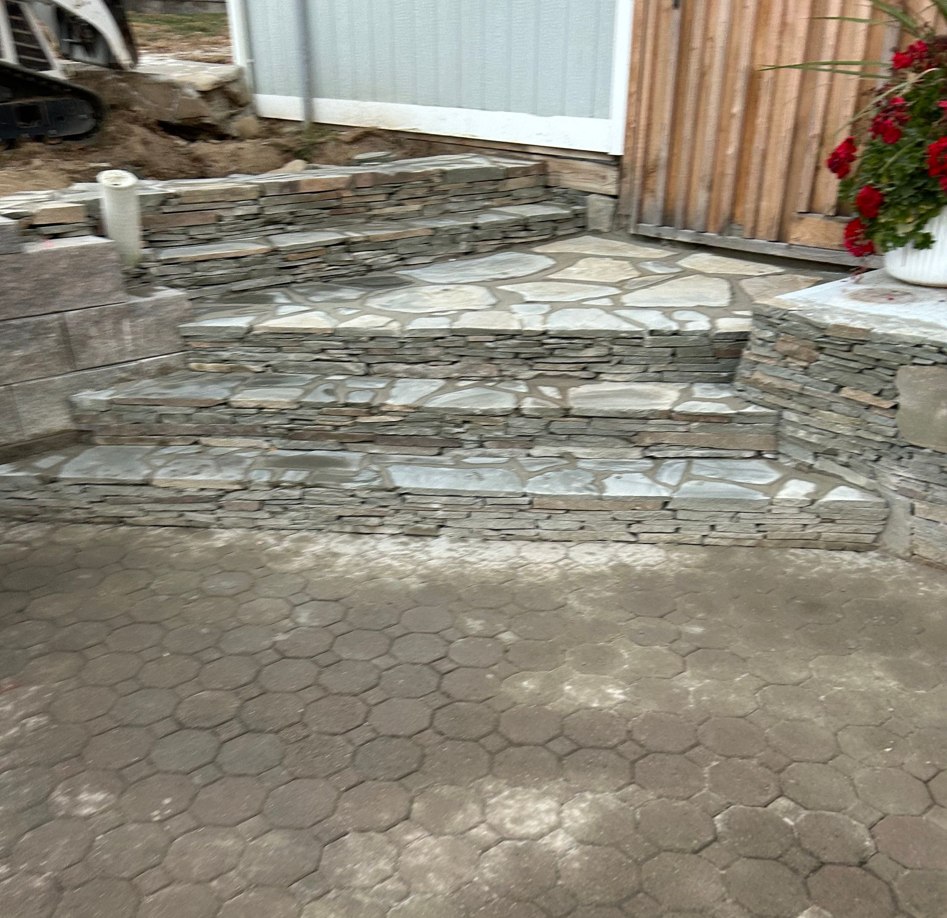 A person using a leaf blower on a patio, shed and fire pit in a yard with a retaining wall and white fence.