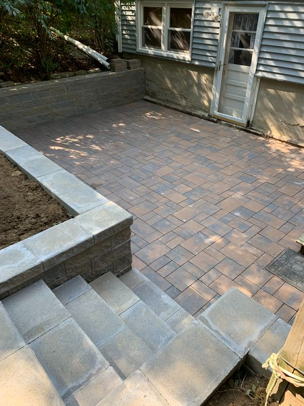 Stone patio with steps and retaining walls near a building with a white door and windows.