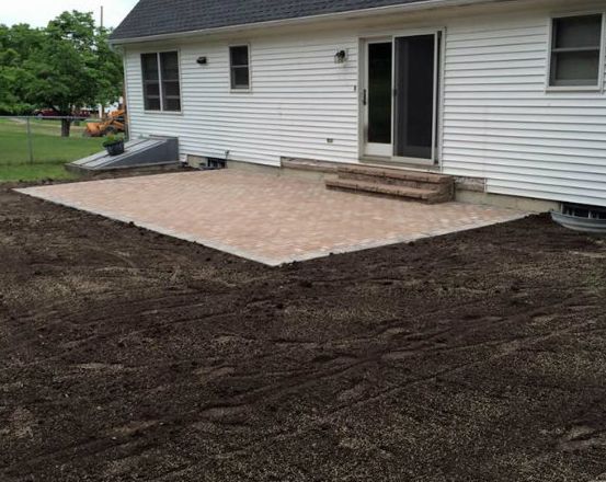Finished paver patio outside a white house with a sliding door and steps. The yard is bare dirt.