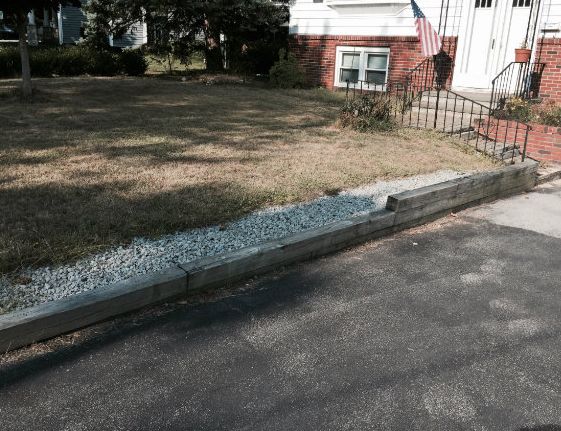 Low wooden retaining wall with gravel and dry grass. Brick house in background.