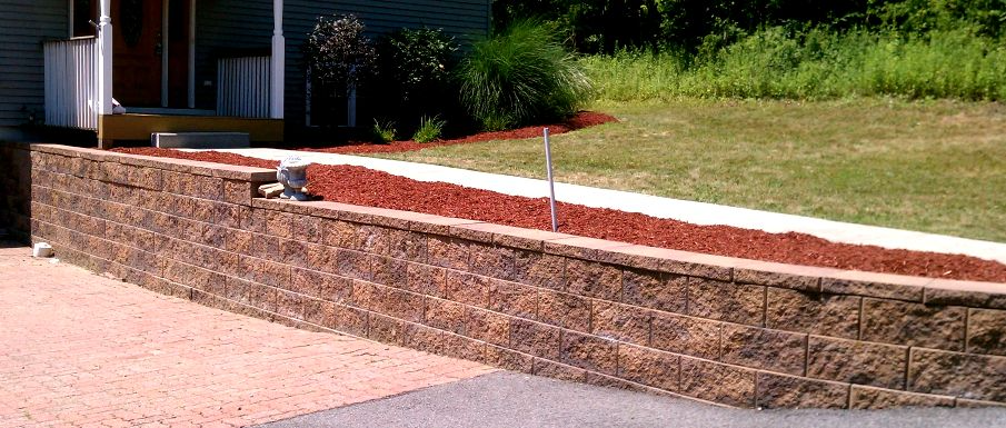 Stone retaining wall with a bed of red mulch, leading to a house. Green grass in the background.