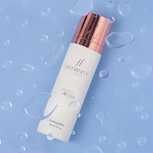 A white and rose gold hydrating mist spray bottle on a light blue background with water droplets.