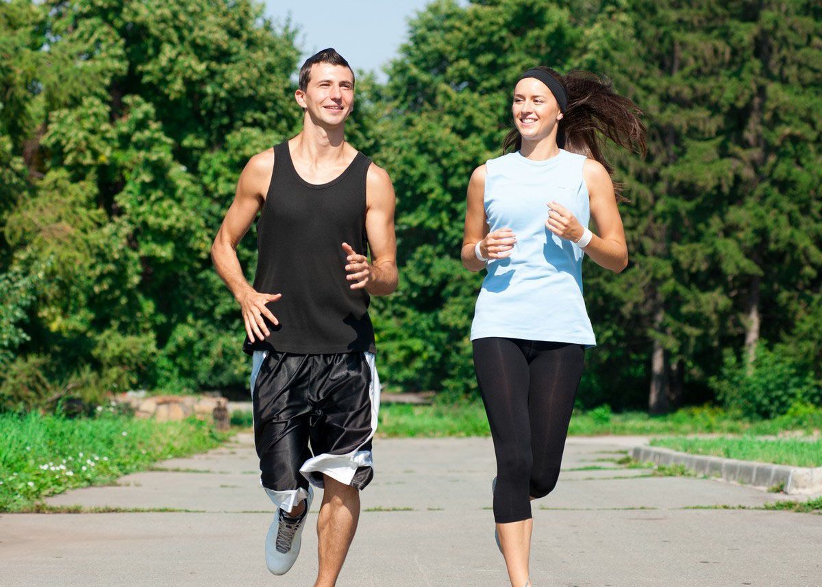Man and woman jogging on a paved path in a park; sunny day, smiles.