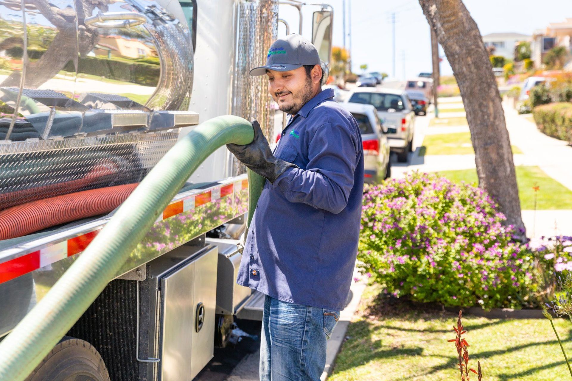 Man in blue shirt, holding a green hose near a septic tank truck on a sunny suburban street.