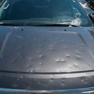 Car hood with many hail dents and water droplets, parked outdoors