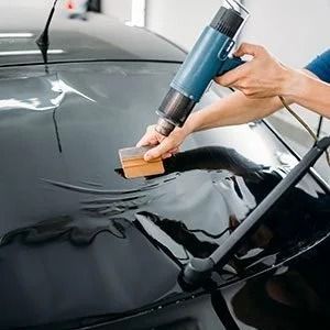 A hand applies black car wrap film with a heat gun and squeegee on a vehicle hood.