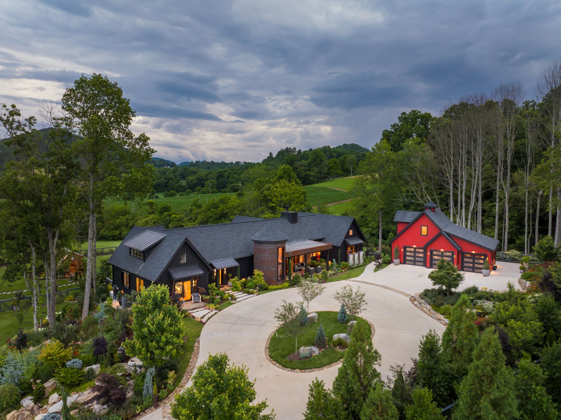 Aerial view of a modern black house and red cabin with landscaped driveway, surrounded by trees and fields