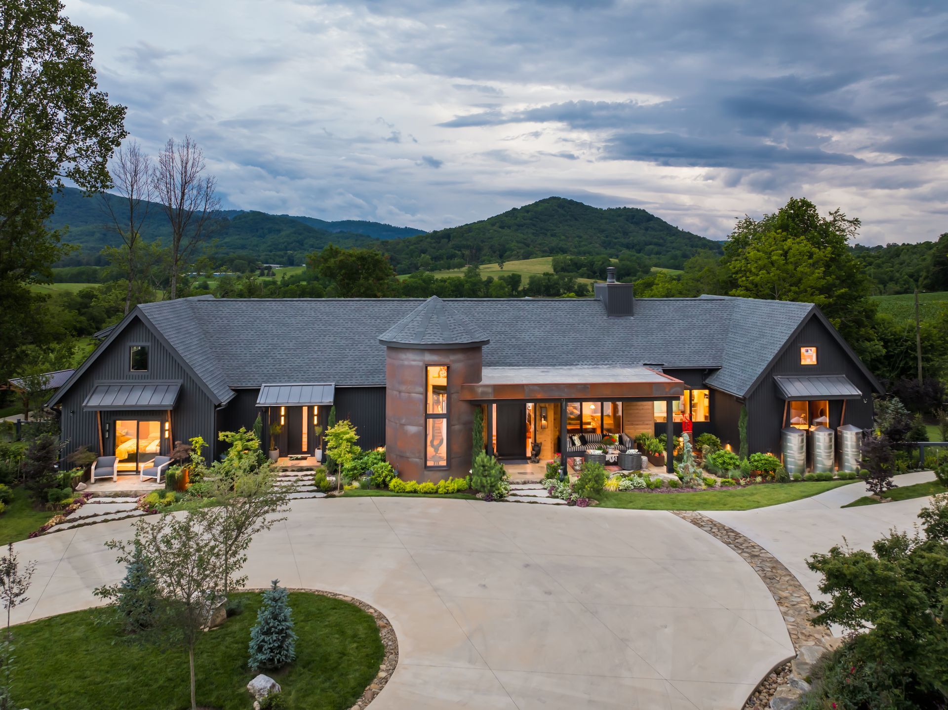 Modern rustic house with lit windows, surrounded by trees and hills at dusk