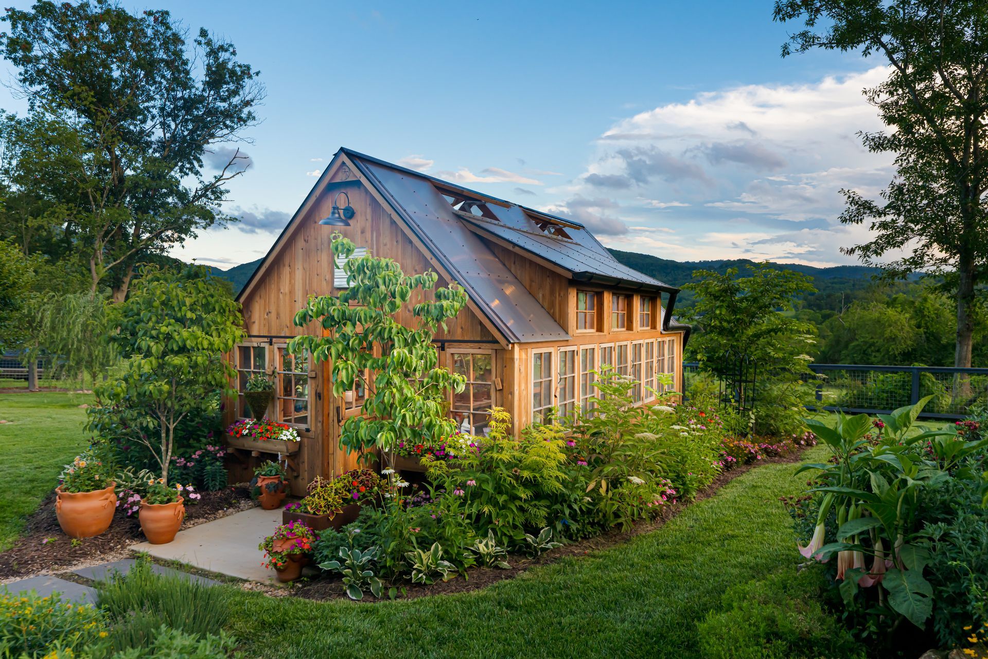 Wooden cottage with a lush garden and potted plants, nestled in a green countryside landscape.