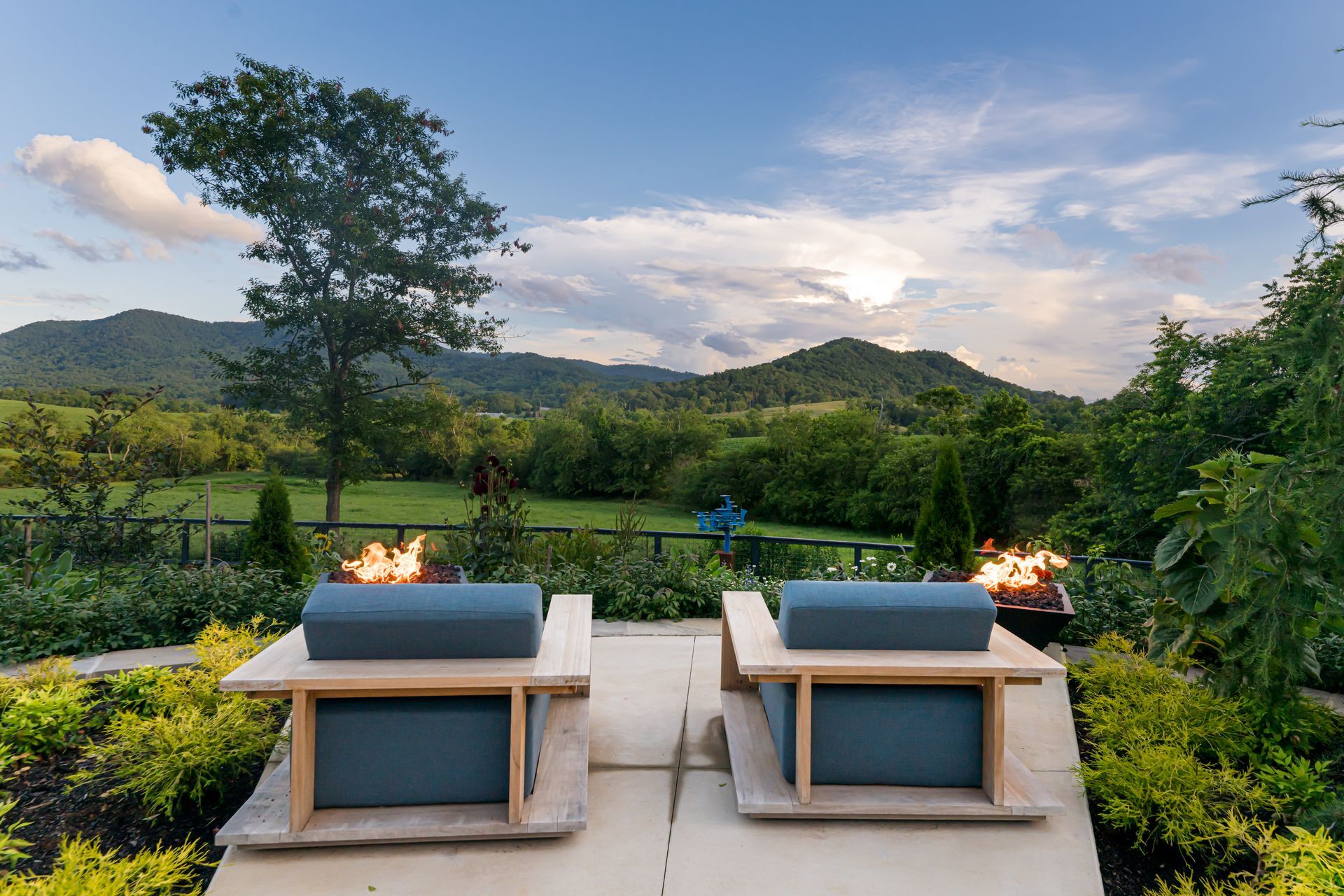 Two blue lounge chairs by a fire pit overlooking green hills at sunset