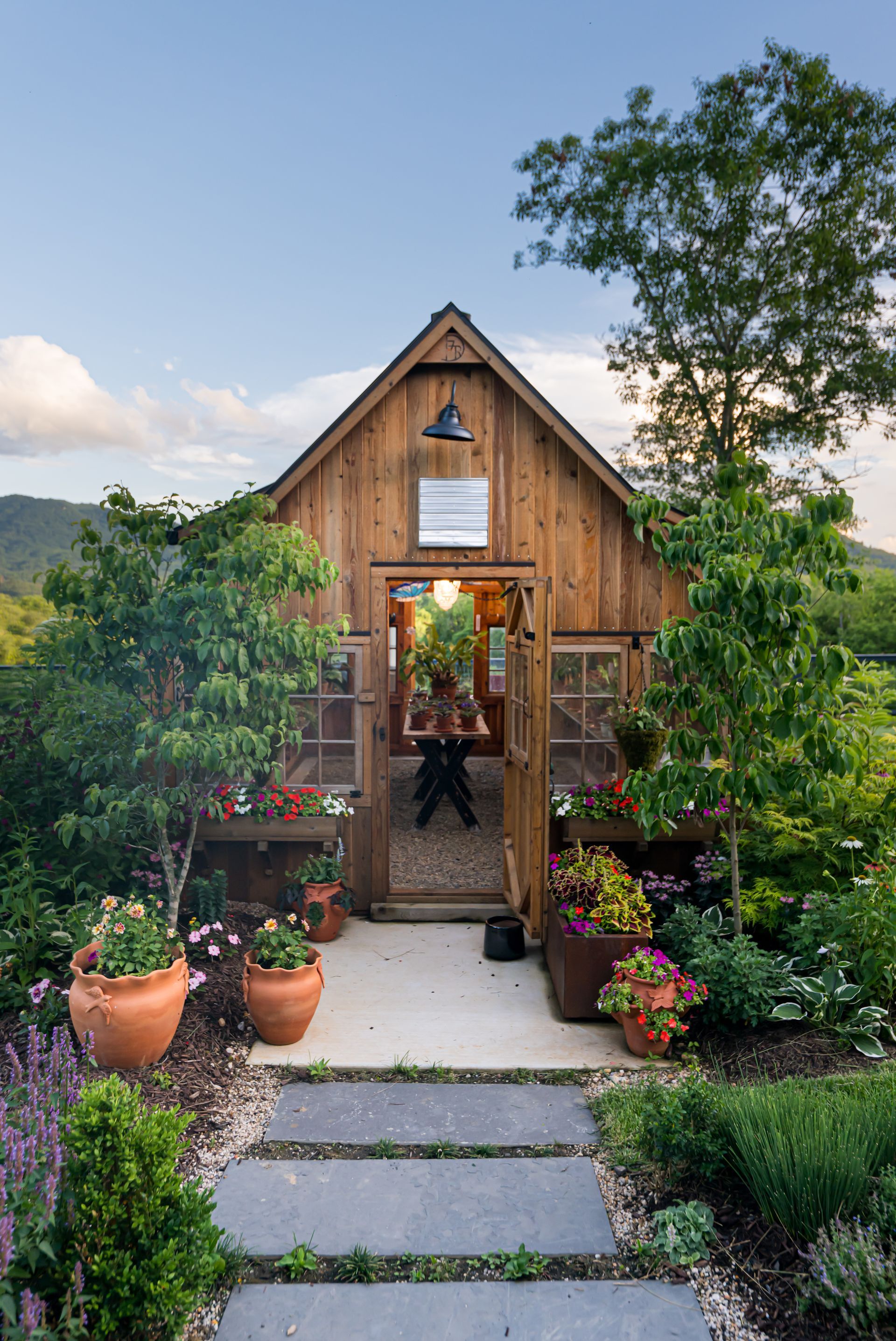 Wooden garden greenhouse with potted flowers and a stone path, surrounded by lush greenery under a blue sky