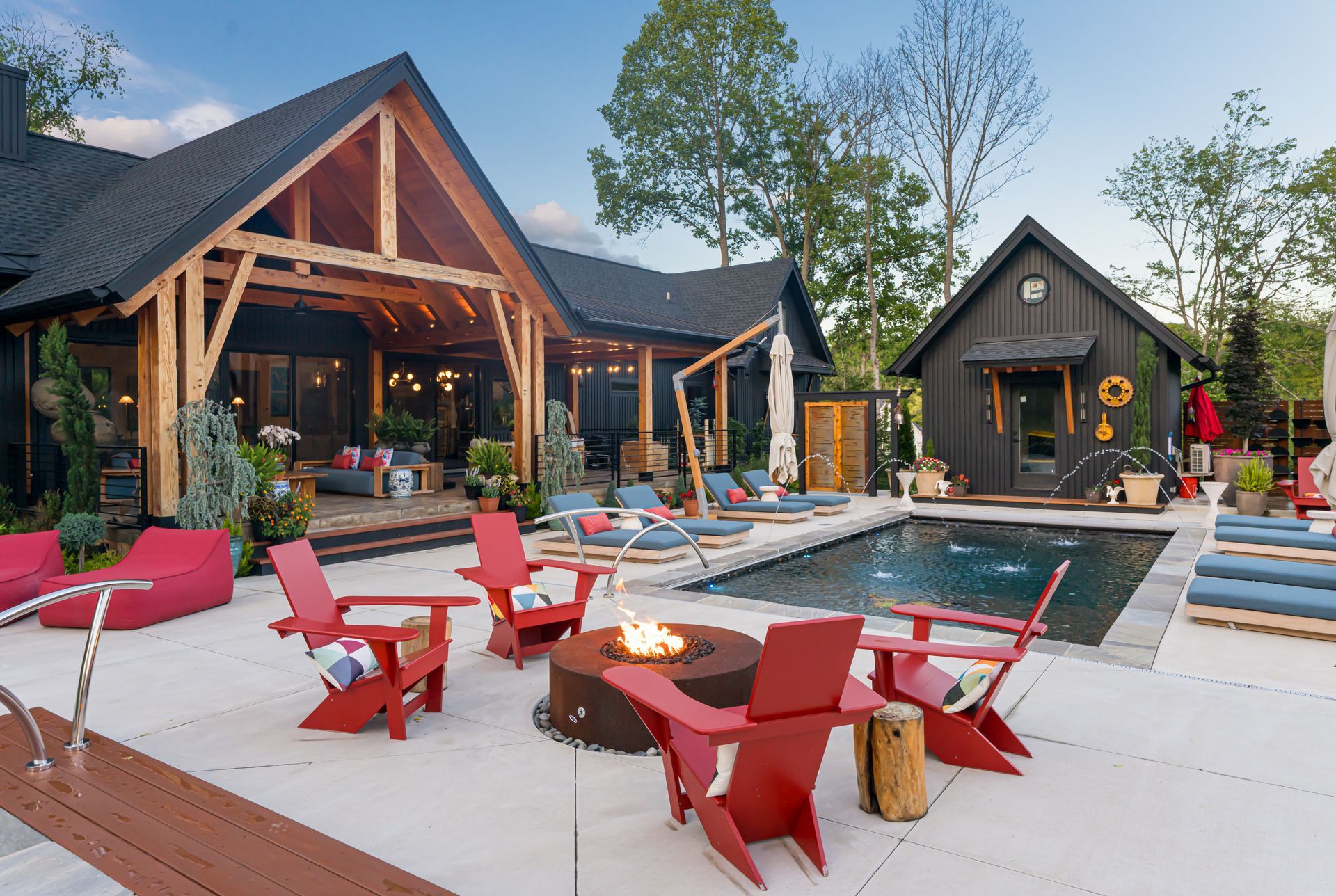 Modern backyard patio with pool, red chairs, and a rustic home at dusk