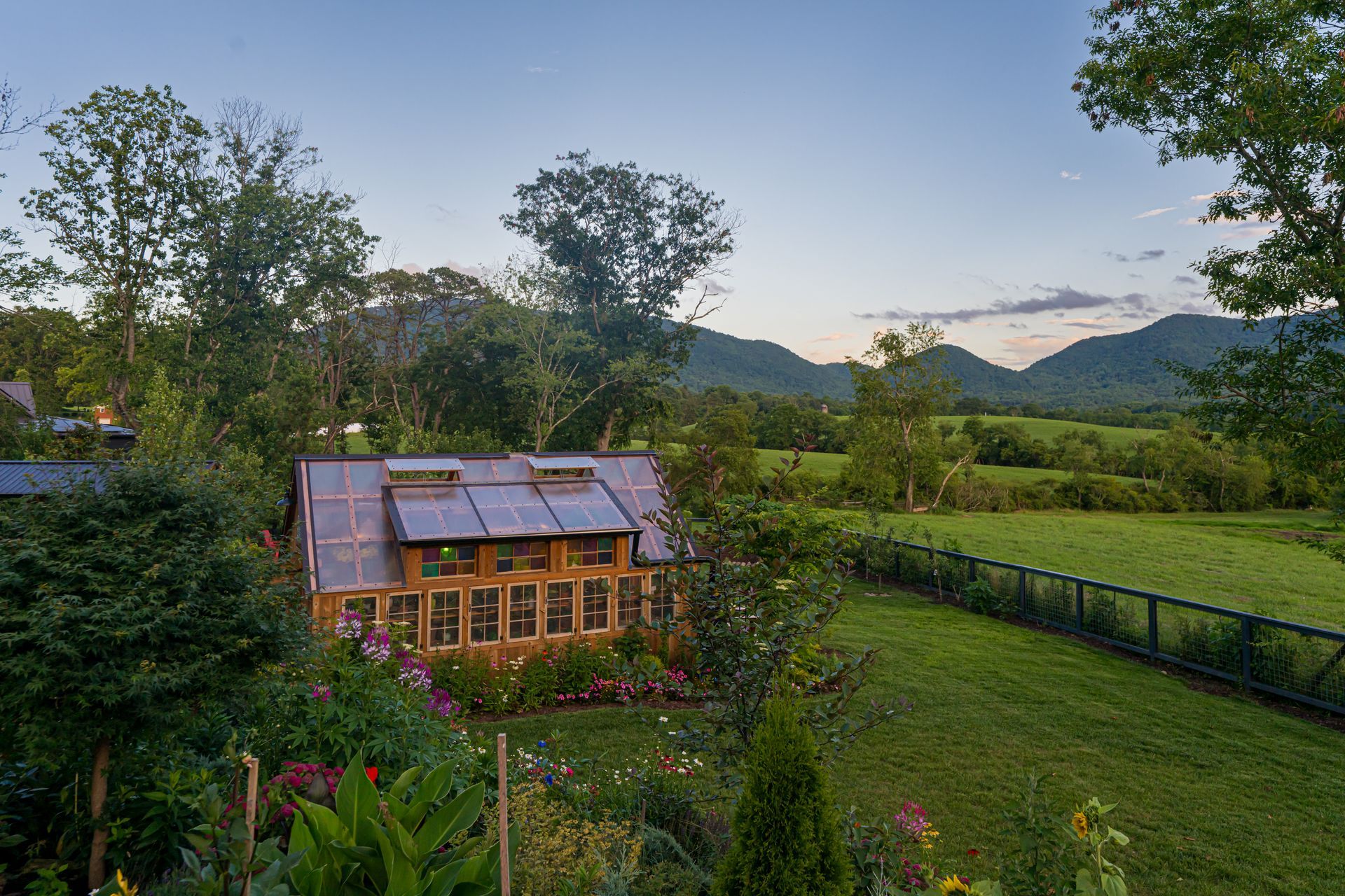 Small rustic cabin in a green meadow at dusk, with trees and mountains in the background.
