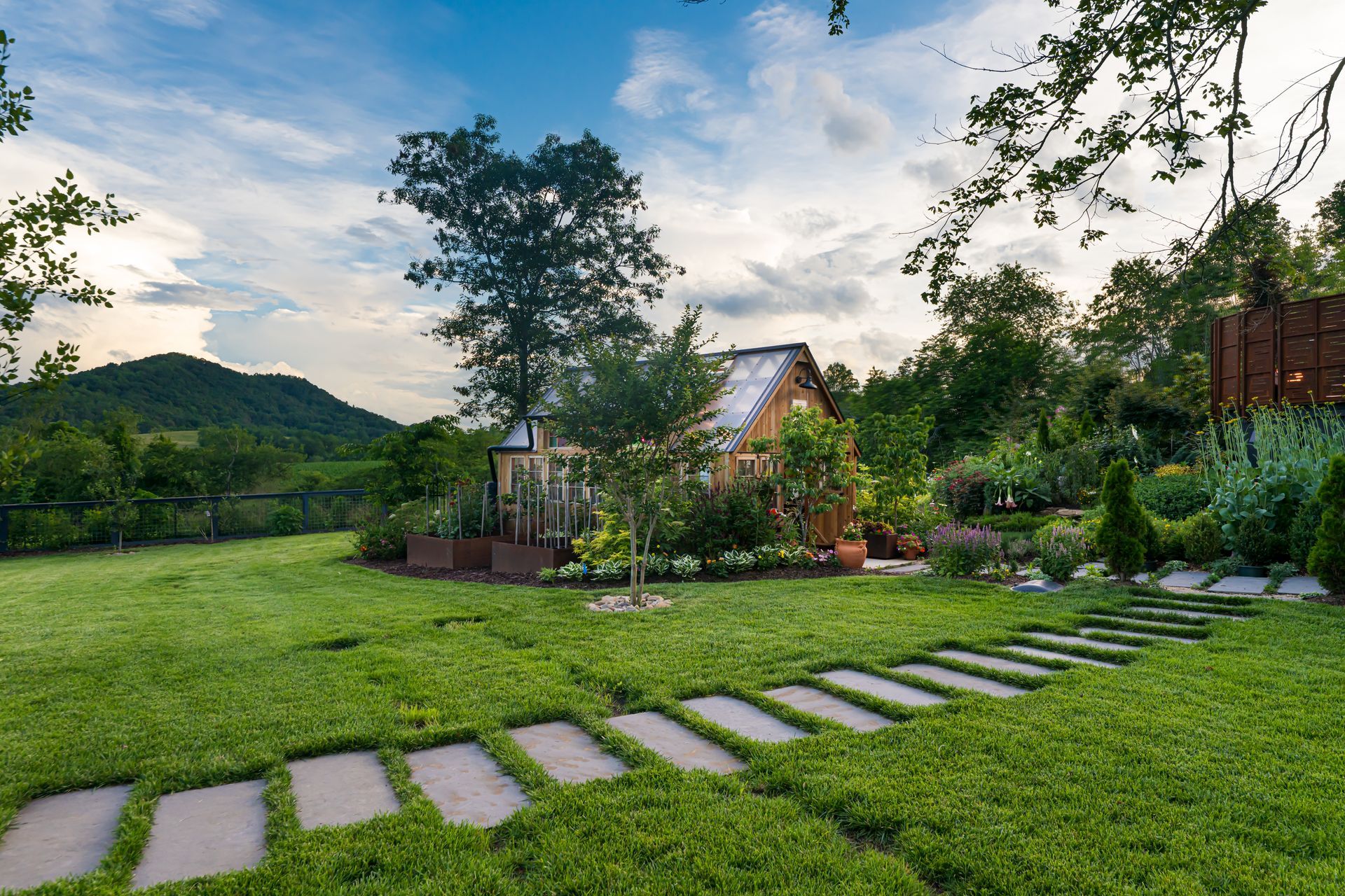 Stone path through a lush green garden with a house, trees, and mountains under a partly cloudy sky