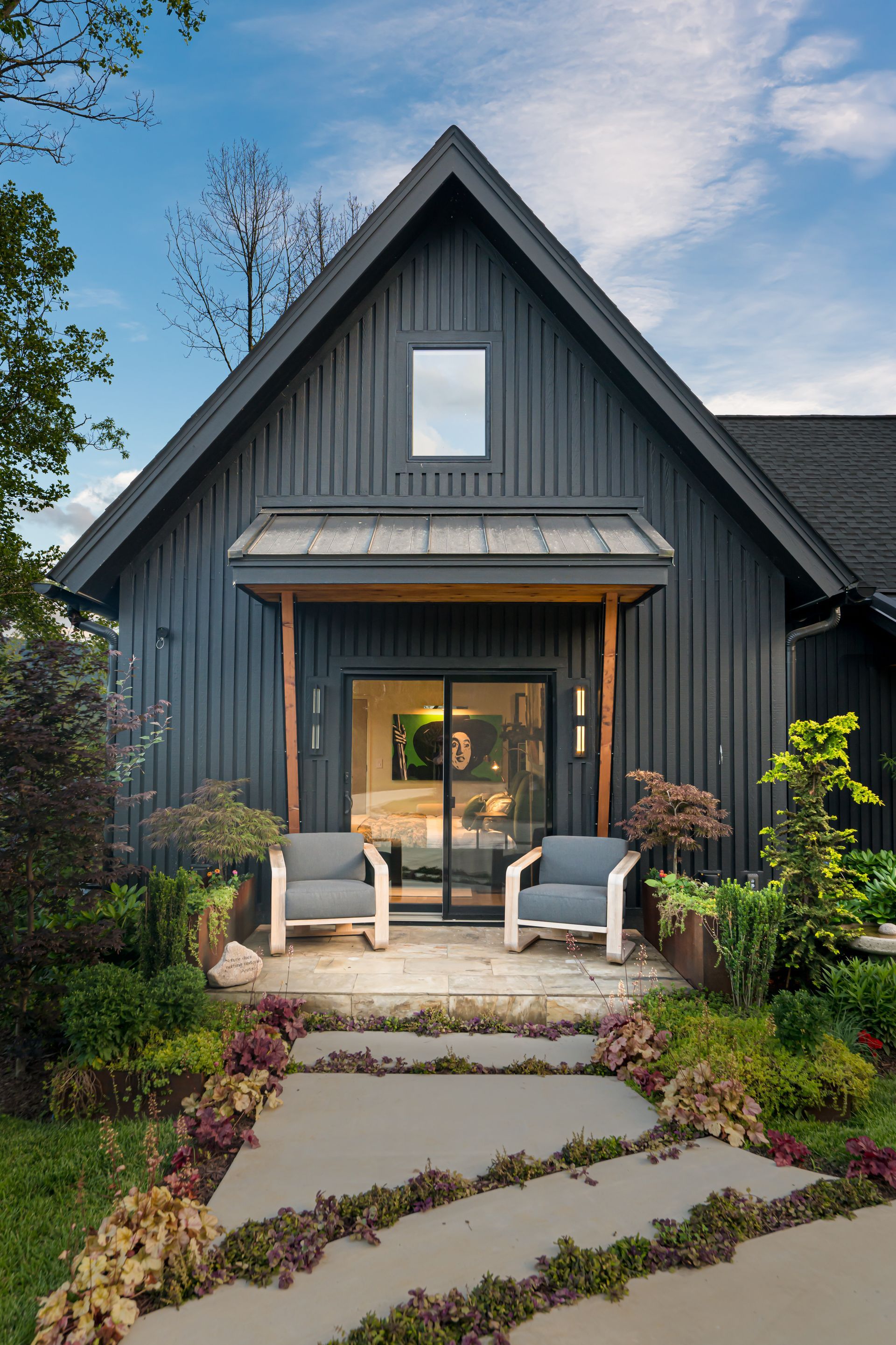 Dark gray cottage with porch chairs, stone path, and landscaped garden under a blue sky