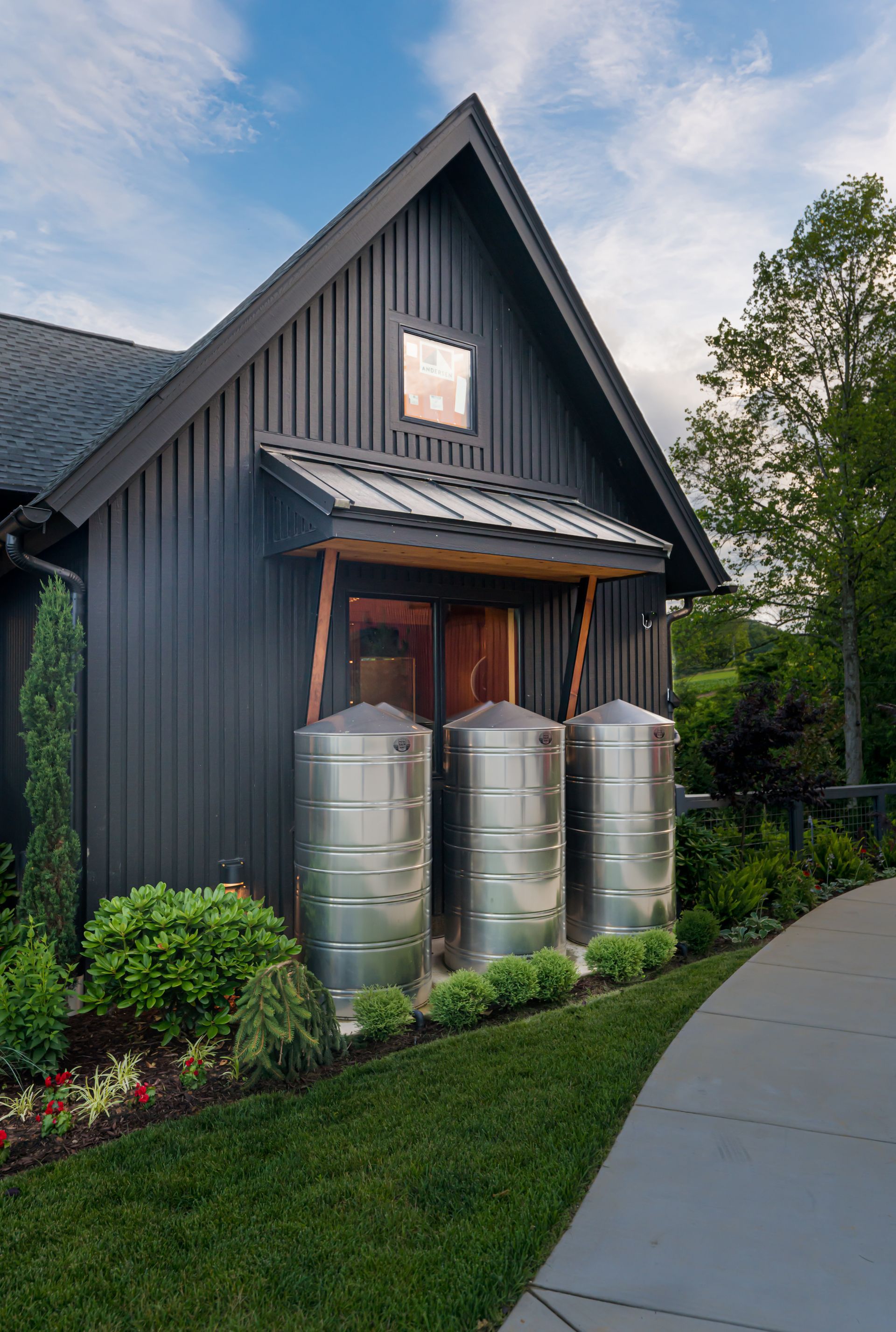 Dark gray house with a steep roof, three metal rain barrels by the door, and a curved sidewalk at dusk.