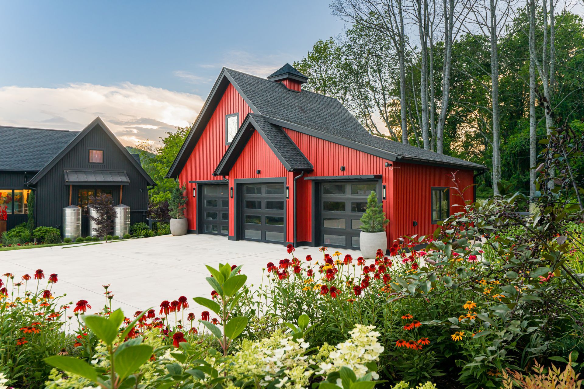 Red house with black garage doors beside a black cabin, surrounded by colorful flowers and tall trees.