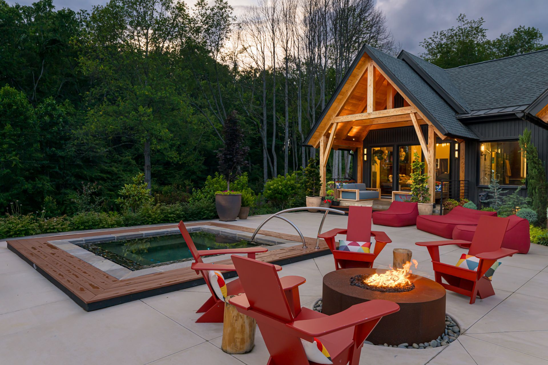 Modern patio with red chairs around a fire pit beside a pool and a lit cabin at dusk
