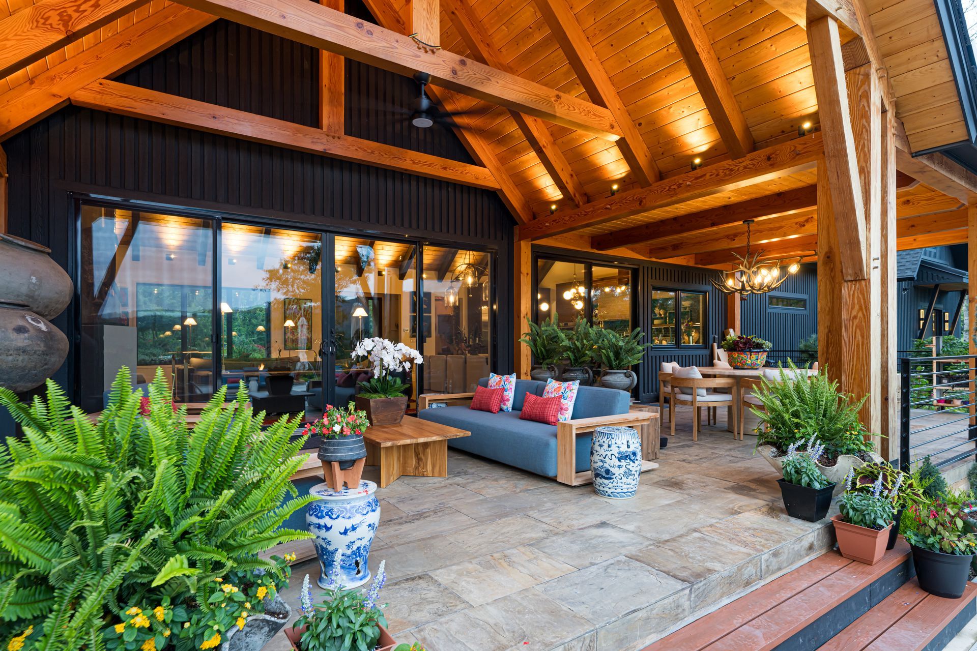 Cozy covered patio with warm wood ceiling, stone floor, blue sofa, dining table, and lush potted plants.